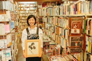 飯田蝶子直筆の色紙をもった笑顔の女性。書店内で。